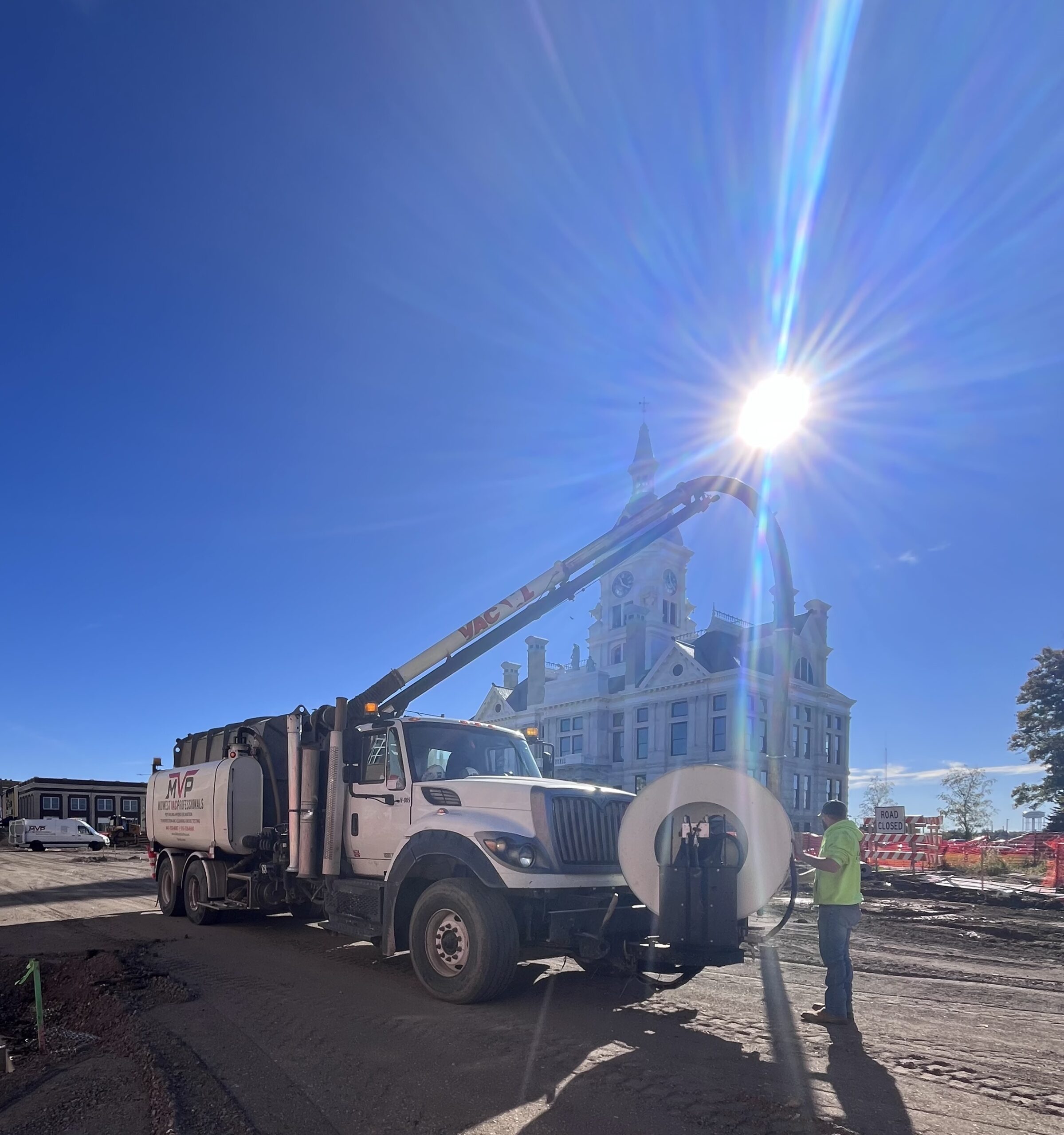 MVP vacuum truck with extended boom hose pumping on a job site near a historic courthouse, with a crew member in a high-visibility shirt operating the equipment.