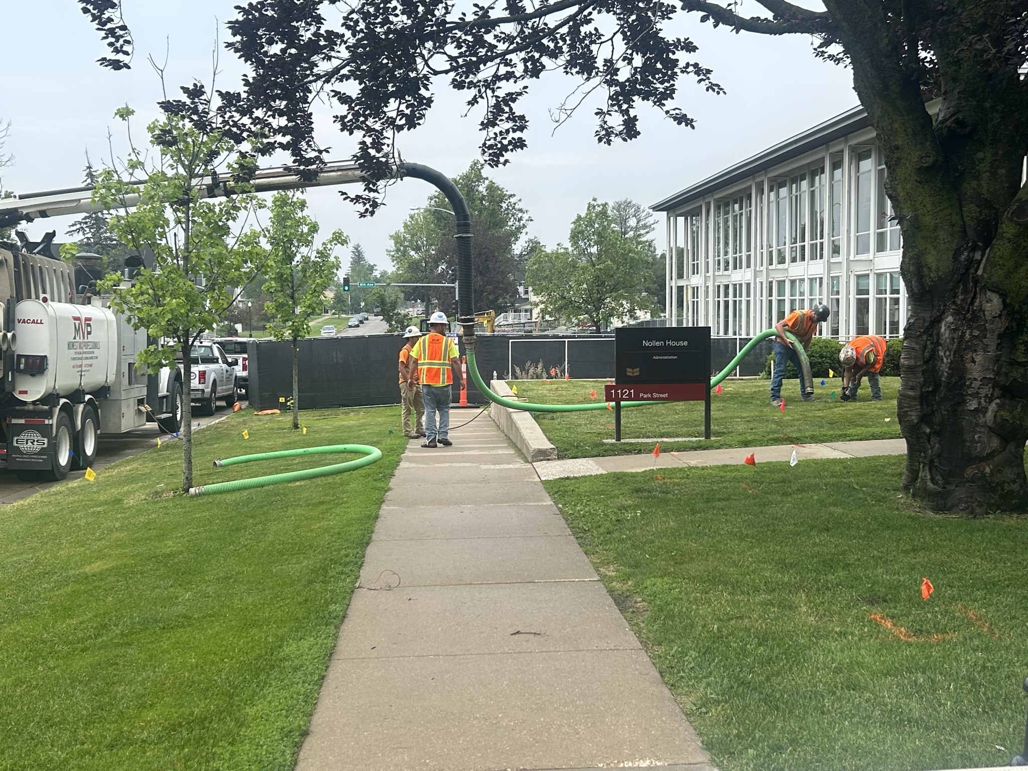 MVP vacuum truck with hose extended across a grassy lawn toward a campus building, with crew members in high-visibility gear working along a sidewalk.